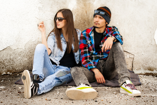 Young hipster guy with his girlfriend sitting in the shadow of an old building. Resting on a hot summer day. Smoke cigarettes and listen to music.