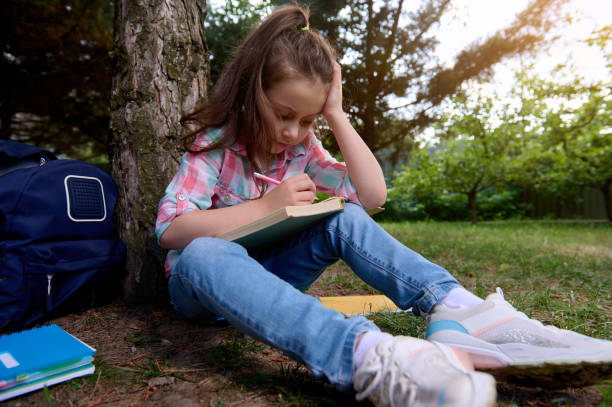 istock Adorable smart little kid girl writing on textbook, reading a book, doing her homework after class in the park at sunset 1587965964