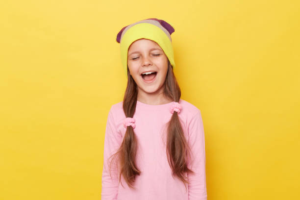 Funny laughing little girl with ponytails wearing pink shirt and beanie hat posing isolated over yellow background standing with closed eyes hearing jokes. Funny laughing little girl with ponytails wearing pink shirt and beanie hat posing isolated over yellow background standing with closed eyes hearing jokes. child laughing hysterically stock pictures, royalty-free photos & images