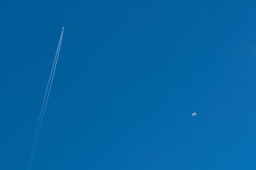 Blue clear sky with a flying airplane and a half-full moon on the opposite side