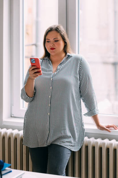 istock Beautiful Plus Size Woman Standing by the Window, Using her Smartphone 1588097173