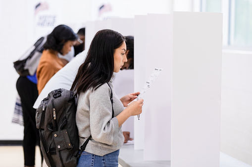 A group of multiracial voters standing in a row at individual voting booths read their ballots before making their choices.