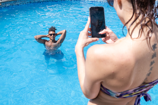 A white woman in a swimsuit takes a picture with her phone of a black man in a swimsuit in a swimming pool, who poses for her.