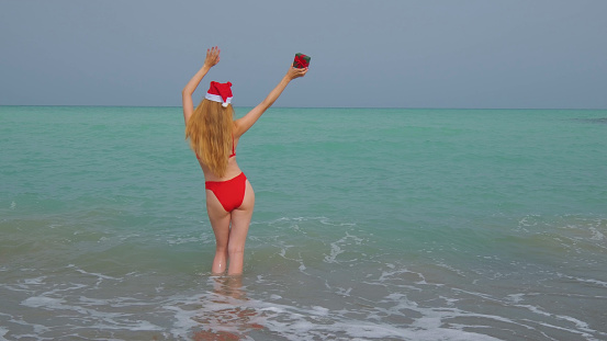 Woman from a back with a long hair, red bikini and in a Santa hat. Mediterranean sea on the background. Blue sky and waves.