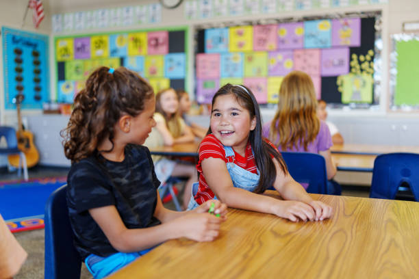 Happy girls talking together at school A cute Eurasian girl sits with a friend at a desk in their classroom and talks cheerfully during class. elementary school stock pictures, royalty-free photos & images