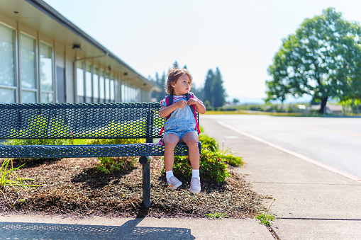 An adorable Eurasian three year old girl wears her backpack and sits on a bench out front of her school while waiting for her parents to arrive and take her home. Independence, education, development and loneliness concepts.