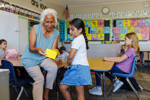 A kind and patient senior adult female teacher of Pacific Islander descent uses flashcards while providing one on one assistance with a female elementary age student during class.