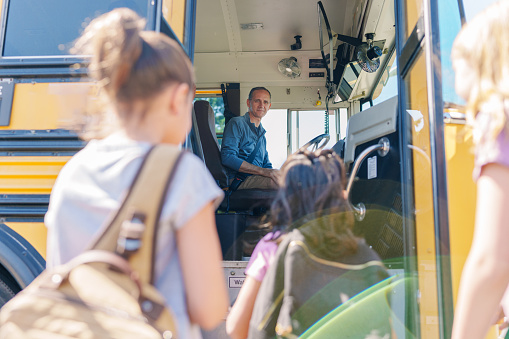 A friendly mid adult man working as a school bus driver sits at the steering wheel of a yellow school bus and greets unrecognizable elementary age kids with a smile as they board the bus.