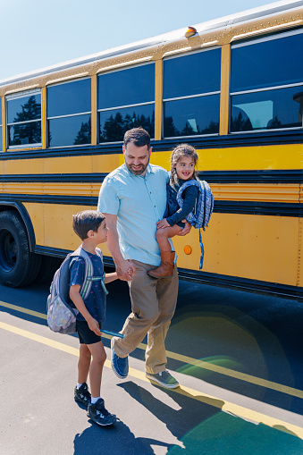 A father carries his cheerful kindergarten age daughter on his hip while walking hand in hand with his son to the school bus stop on a sunny morning. The man is Caucasian and his children are multiracial with Hispanic descent.