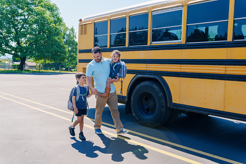 A father carries his kindergarten age daughter on his hip while walking hand in hand with his son to the school bus stop on a sunny morning. The man is Caucasian and his children are multiracial with Hispanic descent.