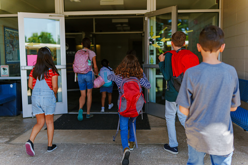 View from outside of a multiracial group of elementary students of varying ages entering their school at the start of the day.
