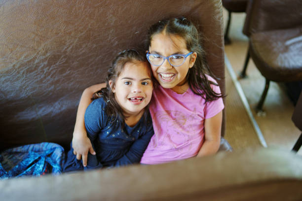 Portrait of a sweet elementary age Hispanic girl affectionately embracing her younger sister who has down syndrome as they ride a school bus together. Both girls are smiling directly at the camera.