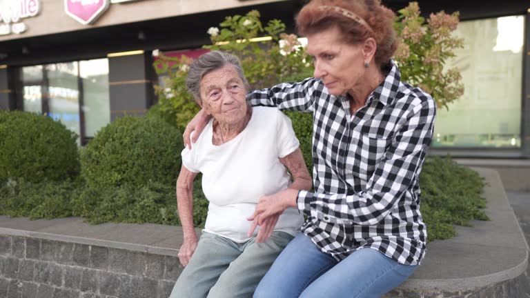 Caring woman with senior citizen walking holding hands and hugging in city. Elderly mom and mature daughter stroll in park by house carefully holding hands and cuddling. Female visiting her old mother