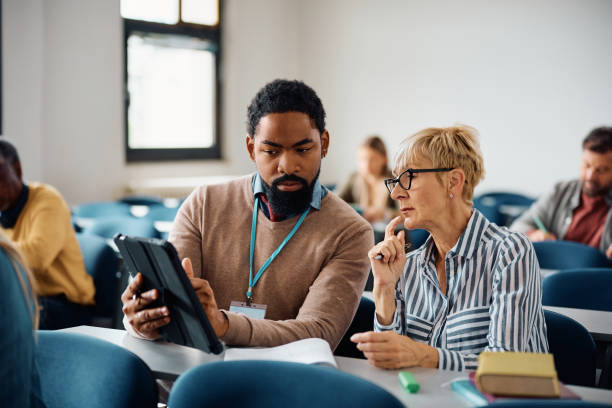 istock Black teacher using touchpad while assisting senior woman with lecture in the classroom. 1588286316