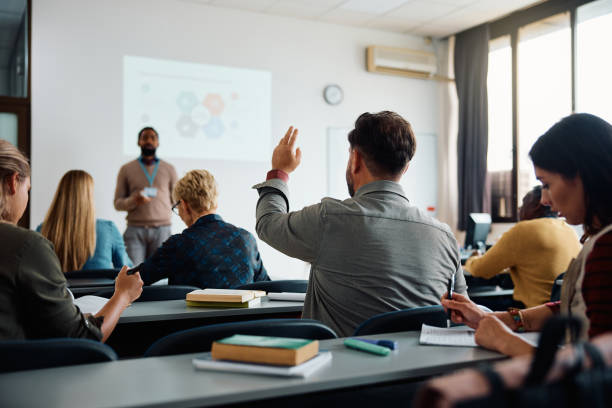 Back view of student raising his hand to answer teacher's question during education training class. Rear view of man raising arm to ask a question during a presentation in lecture hall. mature student stock pictures, royalty-free photos & images