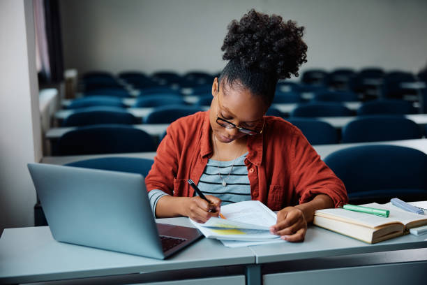 istock Young black woman writing in notebook while e-learning on laptop in lecture hall. 1588291026
