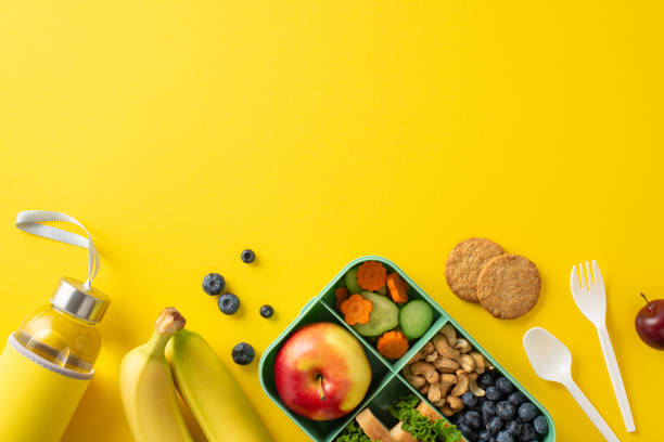 istock Nutritious school lunchbox setup. Overhead shot of eco-friendly container arranged with wholesome snacks, nuts, berries, cookies, water bottle, cutlery on yellow surface with a blank area for text 1588318602