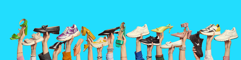 Multiple hands holding trendy shoes of different colors and styles on isolated blue background with copy space. Banner for website header. Shoe store advertising.