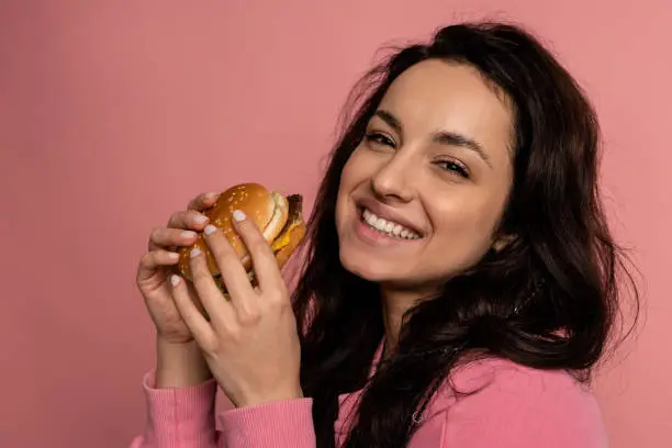 Photo of Pleased brunette with a delicious cheeseburger in her hands smiling during the studio photo shoot. Appetite and favorite fast food concept