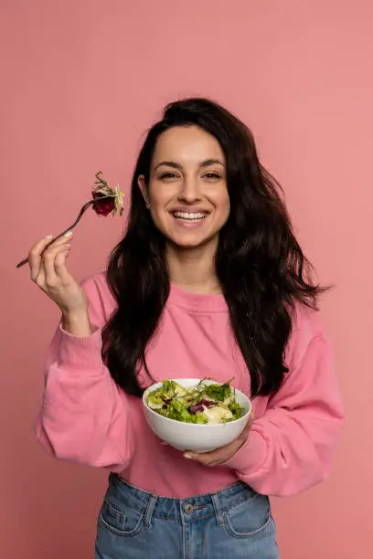 Photo of Smiling happy attractive dark-haired young woman posing on the pink background with her favorite delicious vegetarian dish. Healthy food concept