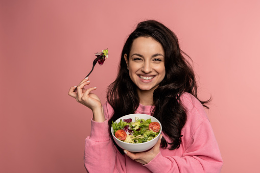Waist-up portrait of a pleased lady eating a portion of an appetizing vegetable salad with the fork from the bowl during the studio photo shoot