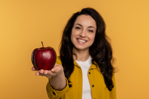 Pleased cute dark-haired young lady holding a red apple on the palm of her hand while standing indoors. Healthy food concept