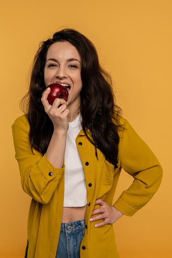 Waist-up portrait of a slim female biting off a piece of an apple against the yellow background. Healthy food concept