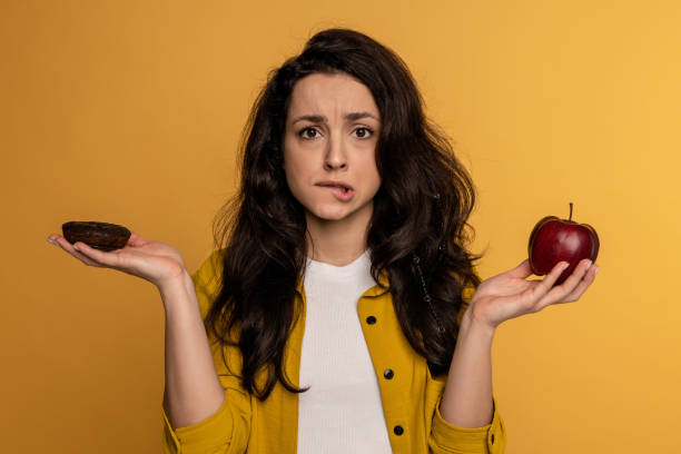 istock Confused dark-haired cute lady choosing between a high-calorie cookie and a low-calorie fruit during the studio photo shoot. Difficult choice concept 1588352149