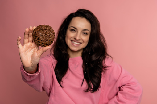 Waist-up portrait of a smiling female holding an oatmeal cookie in her hand in front of the camera. Unhealthy food concept
