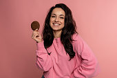 Smiling gorgeous young dark-haired Caucasian woman holding an oatmeal cookie in her hand during the studio photo shoot. Unhealthy food concept