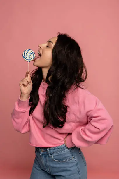 Photo of Attractive brunette licking a hard candy with her eyes closed during the photo shoot in a studio. Sweet tooth concept