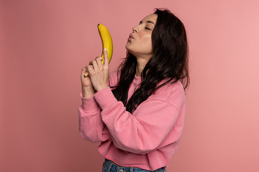 Waist-up portrait of a cute lady blowing at the banana pistol in her hands in front of the camera. Healthy food concept