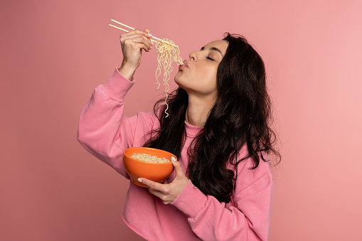 Waist-up portrait of a brunette eating noodles with a pair of chopsticks out of the bowl in front of the camera. Japanese cuisine concept