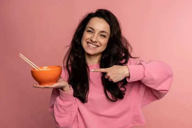 Photo of Smiling friendly cute young dark-haired woman demonstrating her favorite cooked pasta in front of the camera on the pink background. Japanese cuisine concept