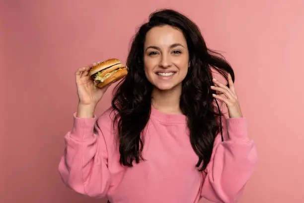 Photo of Joyous dark-haired gorgeous young woman posing on the pink background with her favorite nourishing sandwich. Fast food consumption concept