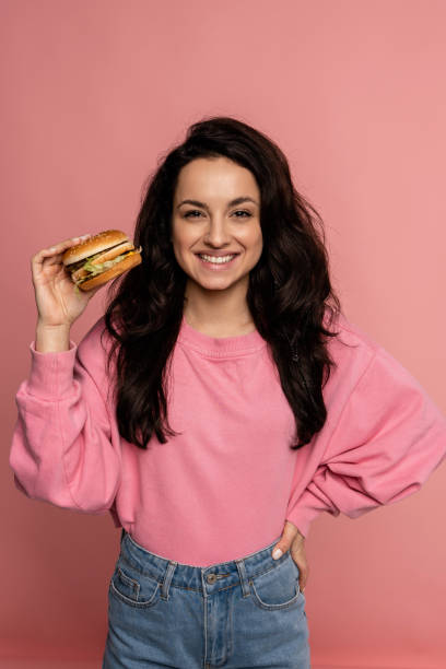 istock Young pretty brunette with a radiant smile showing her favorite nutritious sandwich during the studio photo shoot. Fast food consumption concept 1588354314