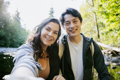 A young couple enjoy a hike through the forest in Washington state, USA.   They take a trail break to look at a river view and take some self portraits with their smartphone.  A relaxing, fun adventure with exercise and beautiful nature scenery.