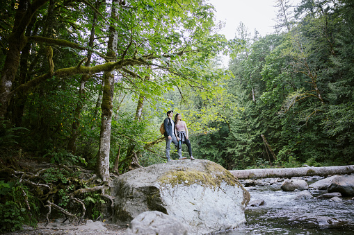 A young couple enjoy a hike through the forest in Washington state, USA.   They take a trail break to look at a river view.  A relaxing, fun adventure with exercise and beautiful nature scenery.