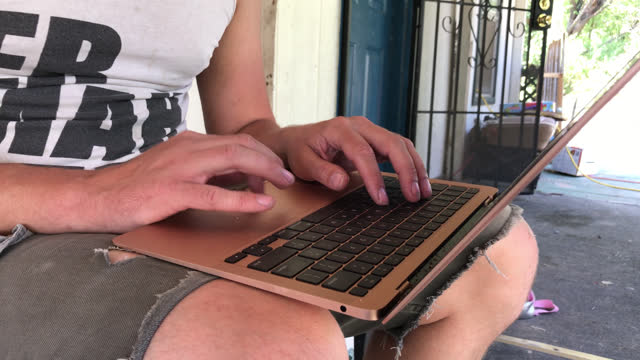 Medium Tight Hand Held Shot Of A White Male In His Early Thirties Wearing  A White Tank Top Shirt Works Remotely On His Laptop Outside A House On A Sunny Autumn Day