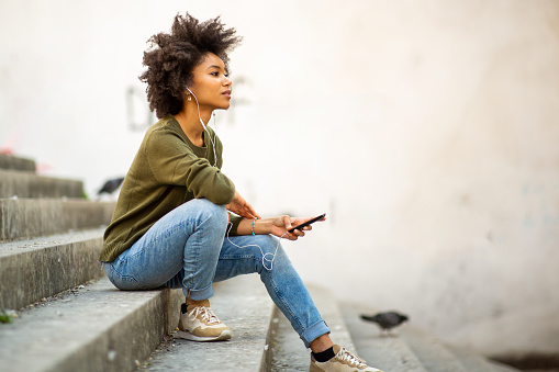 Side portrait of attractive young african woman sitting on steps with mobile phone and wearing earphones