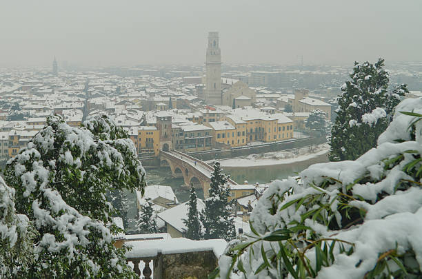 Verona, Italy - Dcember 14 2012: Suggestive morning image of Verona blanketed by the snow and closed in the fog. On the foreground is the river Adige and the old Ponte Pietra, a five-arched bridge and precious testimonial of the Roman period; on the background the imposing Duomo, built on the ruins of two ancient churches destroyed by the earthquake in 1117.The lower part of the tower is Romanesque, the median was built in the '500 by Sanmicheli, while the upper part was continued in the twentieth century by Fagiuoli, but has not been concluded. The tower, 75 meters high, has a set of nine large bells tuned to the scale of La2 waning. It is often cited by European experts campanology as one of the best concerts campanarii existing in the peninsula. These data in hand, one of the largest groups in the world can be played in concert, the bell weighs 4566 kg and has a diameter of 190 cm .