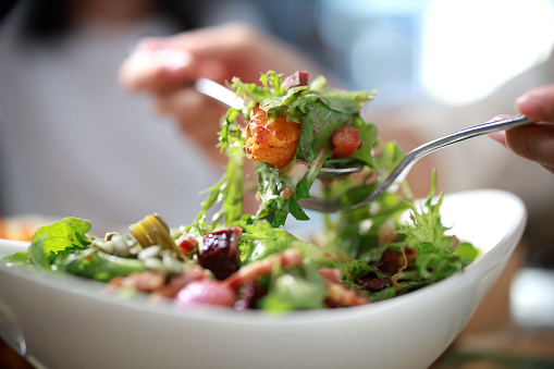 A cropped image of an Asian woman mixing ingredients in her healthy fresh vegan salad. The close-up showcases a multi-colored salad bowl filled with assorted fresh leaf vegetables, including rocket, tomato, zucchini, olives and pumpkin seeds.