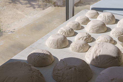 A collection and variety of raw white handmade clay bowls sitting upside down in sunlight next to a large window.