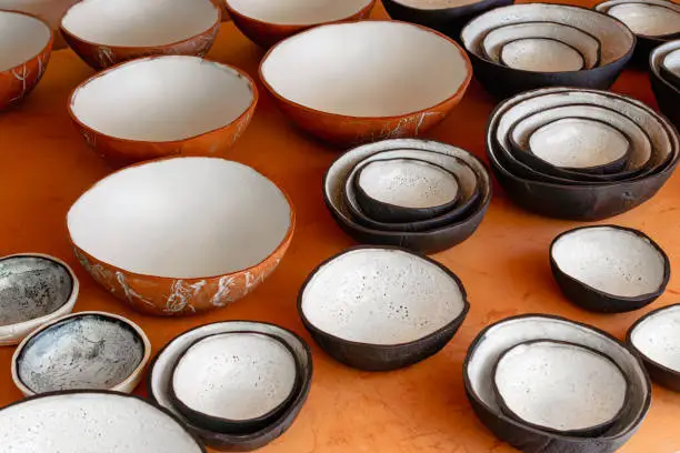A large collection of handmade clay bowls in terra cotta, black, and white glaze in varying sizes. Sitting on a wood table in natural light.