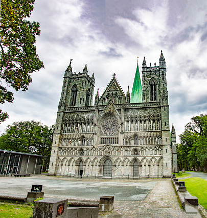 Nidarosdomen gothic cathedral in Trondheim, Norway. Blue and lilac sky with grey clouds, grey medieval stone walls and bell towers, figures of the saints, green grass and trees