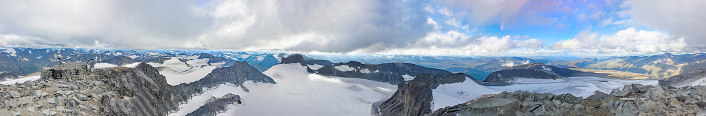 Wide panoramic hilltop view of the mountain landscape of Jotunheimen national park seen from the summit of mount Galdhopiggen in Norway. Blue sky with clouds, grey rocks, white snow