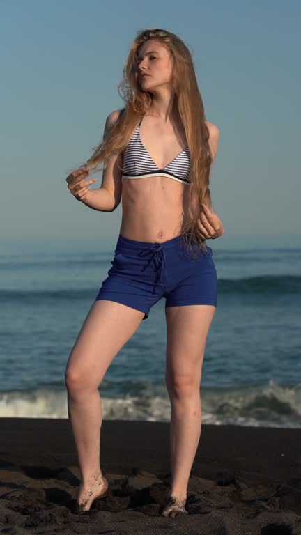 Sensual barefoot woman straightening long hair, standing on beach of Pacific Ocean during summer holidays