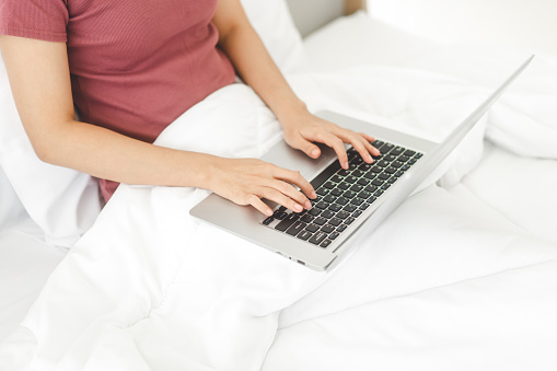 Closeup woman hand typing laptop keyboard on the bed. People using internet for modern lifestyles. Background with white blanket