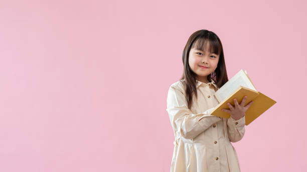 istock A bright Asian girl is holding a book while standing against an isolated pink studio background. 1588962413