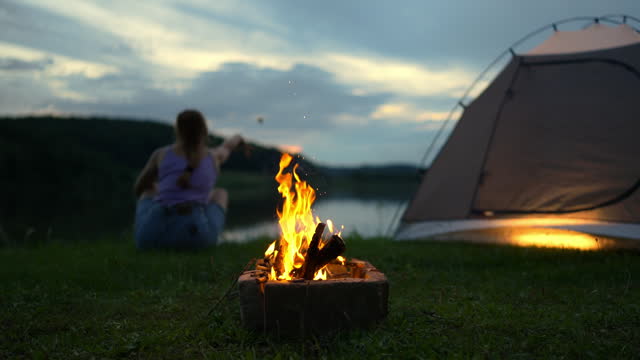 SLO MO Bonfire with Woman Relaxing near Tent and Calm Lake in Countryside at Twilight